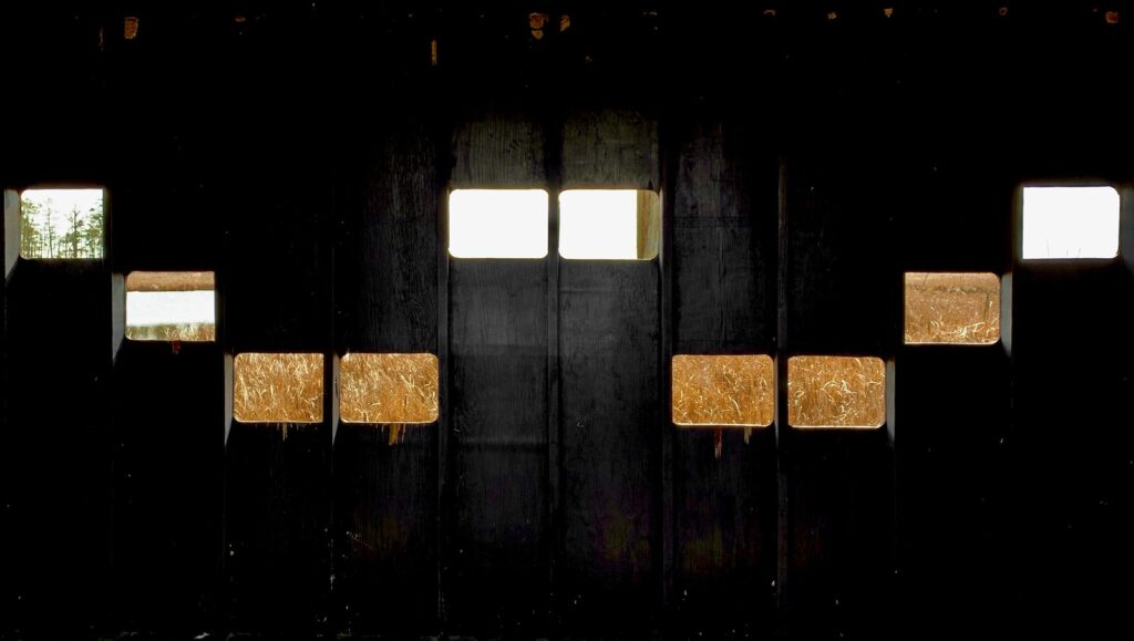 inside of dark barn with a wall with various windows, looking outward; hay bales outside