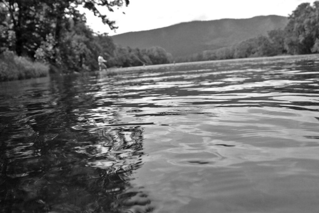 person wearing waders flyfishing in river, focus is on the water with person blurred out