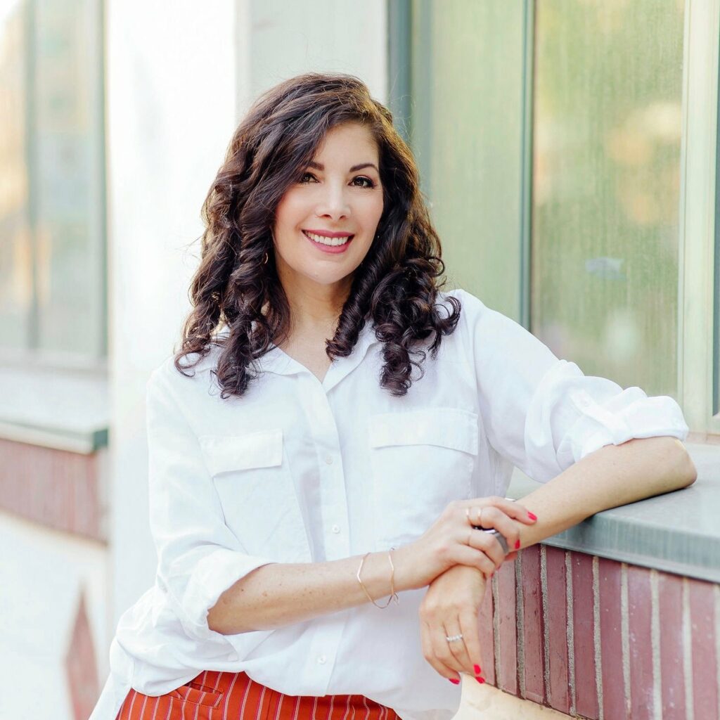 Author Ronit Plank wears a white shirt and is leaning on a railing. She has long dark brown hair and is smiling.