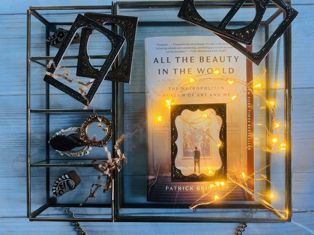 copy of all the beauty in the world by patrick bringley styled on a wooden surface with illuminated strand of lights and old photo frames