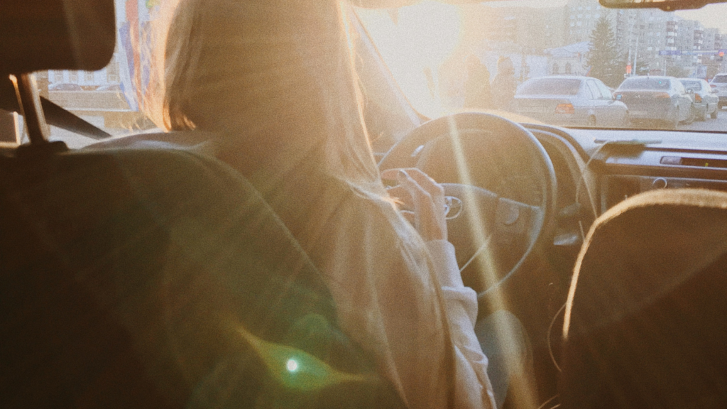 A person holding a steering wheel in city with sun streaming in through windshield