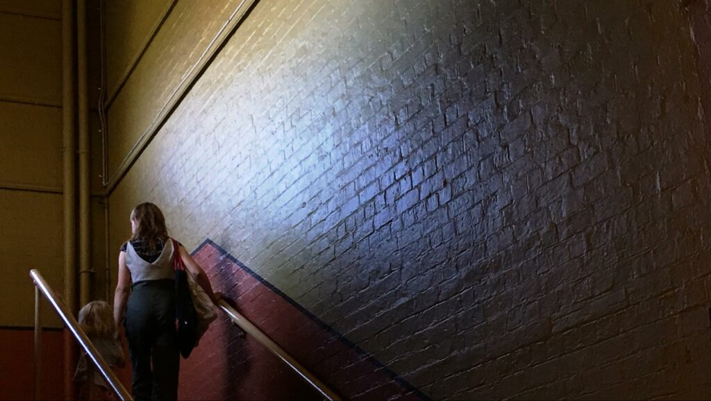woman walking up dark stairwell, concrete walls, bit of sun shining in