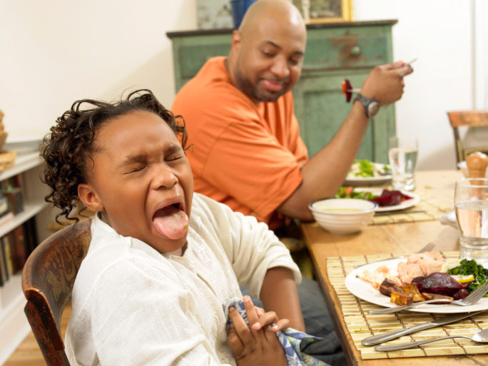 Young Girl Sits at a Table for Lunch With Her Father, Sticking Out Her Tongue in Disgust at the Food