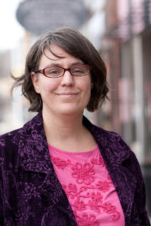 Photo of robyn ryle a white woman with brown chin length hair and glasses wearing a black blazer and bright pink shirt
